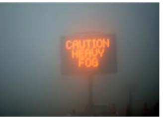 This picture shows a foggy road with a neon sign that reads, 'Caution Heavy Fog.'
