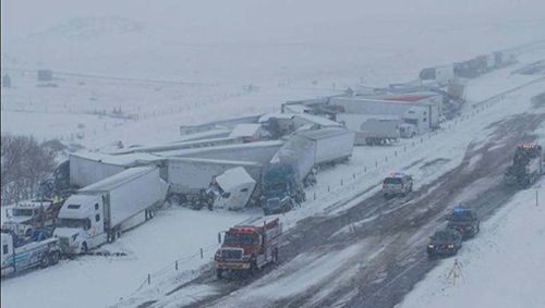 This photo shows a snowy road and background with several large trucks that are on the side of the road. Some of the trucks appear to have crashed into each other.