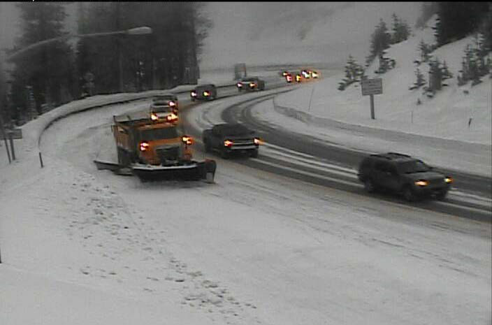 This photo shows a snow plow, trucks, and other vehicles driving on a snowy and curving road.