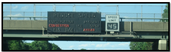 A variable speed limit sign is positioned to the right of a message board mounted on an overpass. The message board indicates congestion ahead.