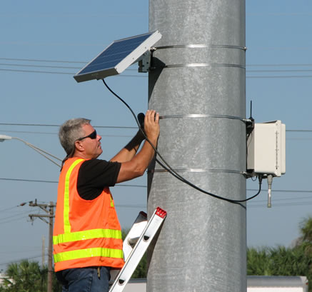 Photo of man in orange vest installing blue tooth device on poll.