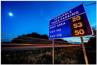 A sign beside a roadway at dusk indicates the availability of truck parking (spaces) at three location ahead.