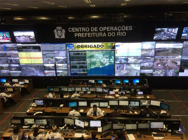 photo of the interior of the Rio Operations Center showing rows of personnel at workstations facing a wall with mutiple screens displaying video feeds, tv news broadcasts, and statistics