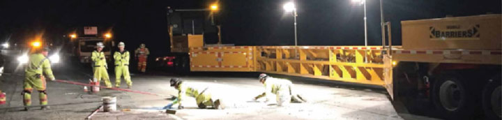 This photo shows a mobile barrier systems set up at night while a maintenance crew of five men work on a roadway.