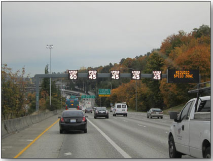 Photograph of the ATM installation along I-5 in Seattle, Washington.  A sign bridge with lane use control signals over each of the 5 lanes is shown with speeds posted on the signs.  A dynanmic message sign is on the right side with 'REDUCED SPEED ZONE' posted on the sign.