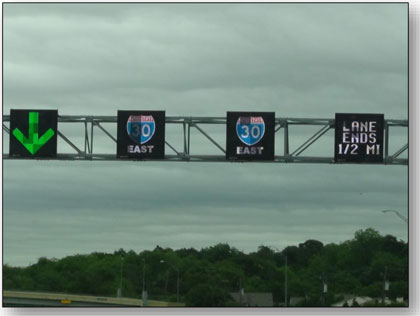 Photograph of the overhead sign bridge used on I-30 in Texas showing dynamic lane use controls.  The left DLUC sign shows a downward green arrow, the two middle DLUC signs show the I-30 East shield, and the right DLUC sign reads LANE ENDS 1/2 MI.