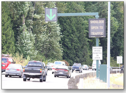 Photograph of the dynamic signing used in Seattle to indicate the shoulder is open to traffic.  A DLUC is illuminated with a green downward arrow over the shoulder and a small DMS on the shoulder reads 'SHOULDER OPEN TO TRAFFIC.'  A statiC regulatory sign reads 'SHOULDER DRIVING PERMITTED ON ARROW.'