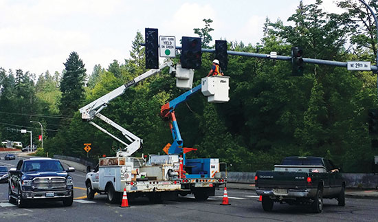 Traffic signal maintenance.  There are two white work trucks with cherry pickers raised. Workmen with orange and yellow vests are working on overhead traffic signals in the middle of the street.