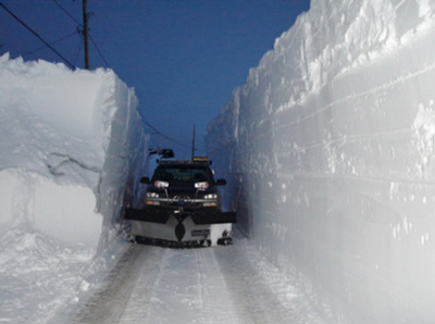 Heavy snow accumulations can make plowing operations challenging. Photograph shows a snow plow working on a roadway with snow banks rising above the vehicle on both sides.