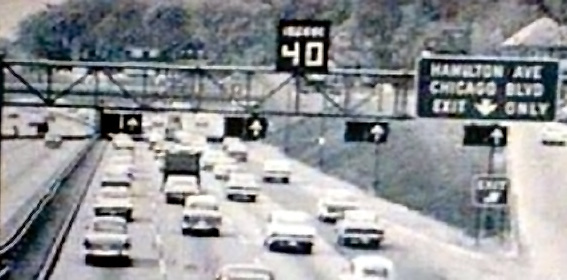 This photograph shows four lanes of the Lodge freeway in the 1960s. There is an electronic sign showing a variable speed limit for all lanes. Over each lane is an electronic sign that can indicate dynamic lane use. The speed limit and lane use signs can be changed electronically to address traffic issues.