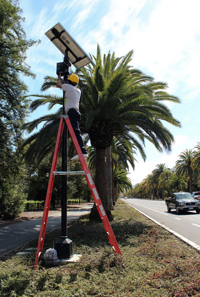 This photograph shows a worker making adjustments to a scanner. The scanner is on a fifteen foot pole located approximately ten feet off a street with passing vehicles.
