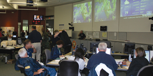 Figure 3 shows a photo of the Florida State Emergency Operations Center in Tallahassee. In the photo, over a dozen people are collaborating on emergency operations and maps of Florida and weather information is being projected onto the wall.