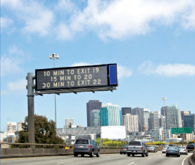 Vehicles heading into a city center on a freeway pass under a permanent, pole-mounted dynamic message sign providing travel time information.