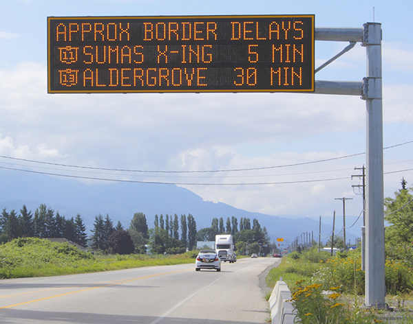 a variable message sign over a rural highway with the message "APPROX BORDER DELAYS | SR 11 SUMAS X-ING 5 MIN | SR 13 ALDERGROVE 30 MIN"