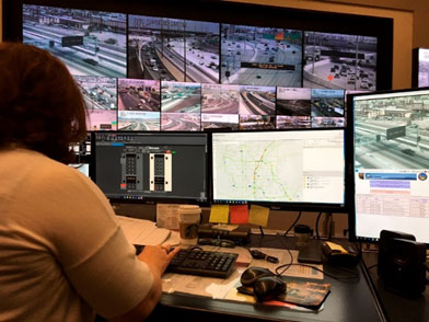 Someone working at a traffic management center workstation that has 3 computer monitors, facing a wall of videos, tracking what is happening on the freeway and arterial system.