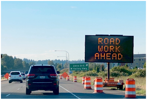 A dynamic message sign at the shoulder of a roadway warning drivers of road work ahead.