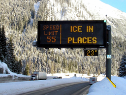 A permanent, pole-mounted dynamic message sign warning over a snowy roadway that warns drivers that there is ice in places.