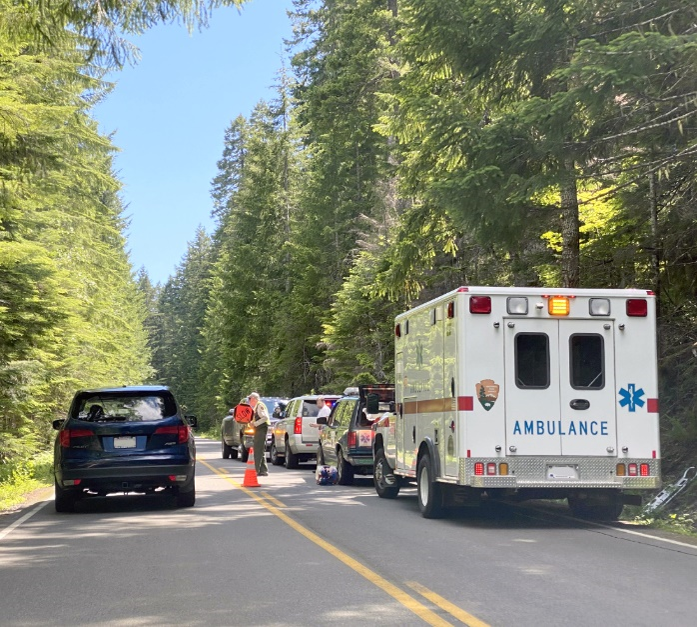 An ambulance and a line of cars on the right side of a two-lane rural roadway with trees on both sides. A person with a paddle that reads 'Slow' is assisting cars in passing the stopped vehicles.