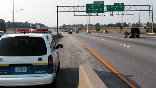 photo showing automobiles in three lanes of a divided highway, with a traffic control vehicle parked on the left shoulder, orange cones in the gore between the right lane and the exit ramp blocking the exit, and another traffic control vehicle parked on the closed exit ramp facing oncoming traffic