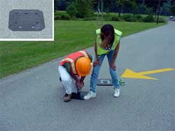 photo of two technicians installing a small electronic vehicle counter on a road surface. An inset photo shows the counter as a flat black square with holes and slits around the outside edges