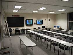 photo of a meeting room with a flip chart and desk in front of three long tables with chairs; wall-mounted monitors and computer monitors also are shown
