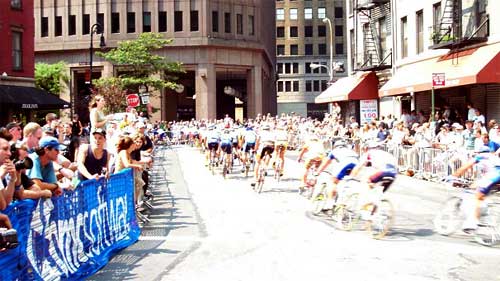 photo of a group of bicyclists racing on a street closed to traffic by barricades on both sides and spectators crowding into the barricades