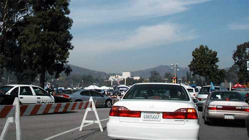 several automobiles traversing three lanes that connect to a Rose Bowl access road during event ingress with a police cruiser parked in the intersection channelization gore area