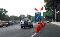 a photo showing several traffic cones extending along a barrier line between the right lane of a freeway and an exit ramp auxiliary lane, and a pedestal-mounted guide sign, erected at the first cone in the upstream direction, stating "general parking – exit 9E"
