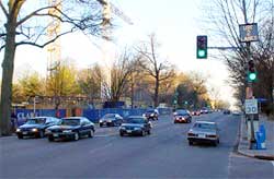 photo showing a four-lane roadway with a center left-turn lane operating as three lanes in one direction and two lanes in the opposite direction, and a dynamic blank-out sign, mounted on a light post at an intersection, displaying the message "use 2 lanes"