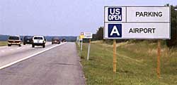photo showing a guide sign, located on the right shoulder of a freeway, displaying a logo entitled "US Open" to the left of the word "parking" followed below by a logo entitled "A" to the left of the word "airport"