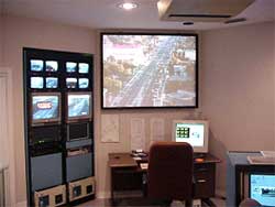 photo showing room containing a desk and computer located in front of a large video screen showing a live traffic scene. A panel of ten smaller video screens and hardware is located to the left of the desk