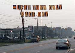 photo showing a sign, hanging over a four-lane roadway, displaying the message "left turn only – race day"