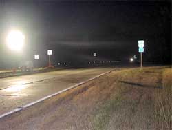 photo showing a device, located in the median of a four-lane road at a freeway interchange, shining a bright light during nighttime conditions