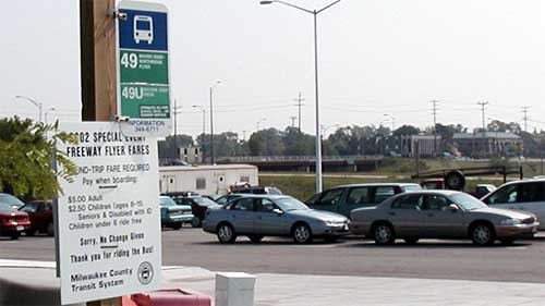 photo showing two express bus signs next to a travel lane in a parking lot filled with cars. One sign is shown as a bus route sign with numbered bus routes, and the other lists bus fares for the 2002 Special Event Freeway Flyer