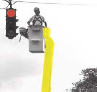 Image - Maintenance worker in a bucket truck being lifted up to a traffic signal.  It appears as though the maintenance worker is going to perform some type of maintenance on the signal.