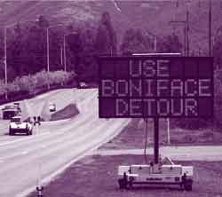 Two cars travel in opposite directions on a highway. A large construction sign on the side of the road warns drivers to use a detour.