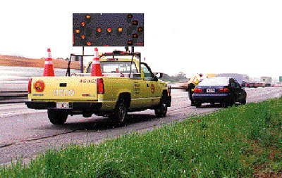 A pickup truck with a mounted warning arrow panel parked behind a car on a roadway right shoulder