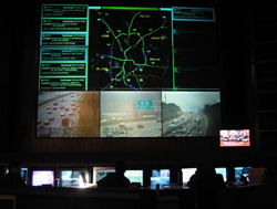 Photo of a Traffic Management Center, showing workers looking at multiple large-screen monitors