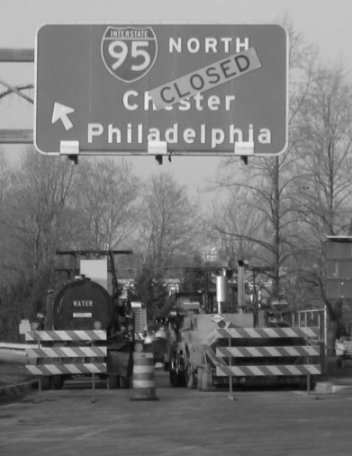 Crews work during a rehabilitation of Interstate 95 near Wilmington, Delaware.  The picture shows an exit ramp with a large overhead sign that reads: Interstate 95 North Chester Philadelphia. The sign has a large yellow placard pasted at an angle across the sign that reads: Closed. In the foreground, a road construction machine and a water truck sit parked behind two safety barricades and a safety cone. 