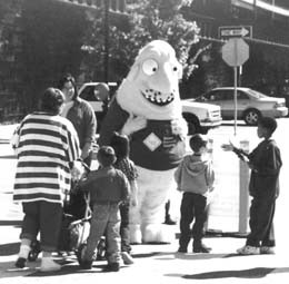 Figure 2a – DelDOT used a character known as the traffic "Creep" in public outreach efforts. This picture exhibits a view of a group of citizens talking with Creep. The Creep is about seven feet tall with a round body and cone–shaped head with no hair. The character's eyes and nose are prominent and he sports a stubble moustache. The scene depicts several children with their mothers interacting with the Creep on a street corner.