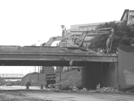 Figure 2 - The requirement for significant bridge repairs made full road closure an attractive option during the M-10. The figure shows a backhoe tearing into the top of a bridge. Under the bridge, the scene shows several pieces of Work Zone equipment.
