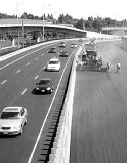 Crews work without interruption on I-84 (Banfield 
Freeway), physically separated from traffic. The picture shows a line of barricades physically separating the workers on the left from the three lanes of oncoming traffic on the right. In the middle of the right hand side of the picture, a pavement grader applies a treatment to the road attended by half a dozen workers.