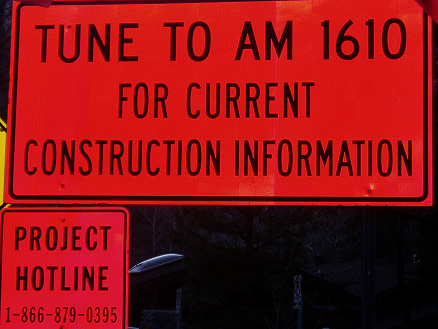 fluorescent pink signs stating: Tune to AM 1610 for current construction information, and Project Hotline, 1-866-879-0395