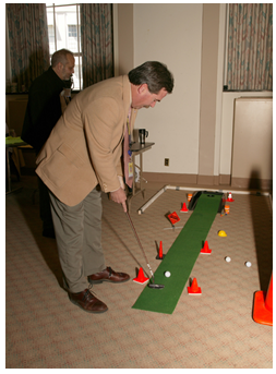 A man playing 'mini-golf' with miniature traffic cones and work zone signs lining the 'green' at a work zone safety awareness event.