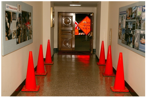 Orange cones lining a corridor in a building. Beyond a set of double doors at the end of the hall are work zone signs that read 'stop' and 'road work ahead'