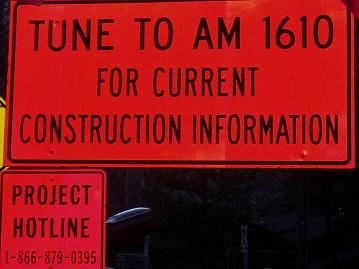 Fluorescent orange road signs stating "Tune to AM 1610 for current construction information" and "Project Hotline, 1-866-879-0395"