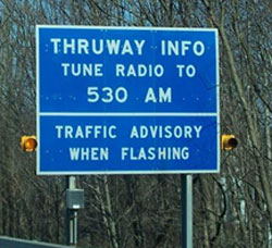 Photo of a blue sign with a warning beacon on each side stating Thruway Info, Tune Radio to 530 AM, Traffic Advisory When Flashing.