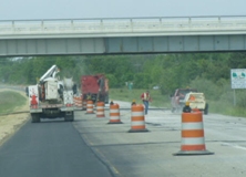 Photo of a road widening project featuring a work truck on the newly paved shoulder separated from through traffic by a line of traffic barrels.