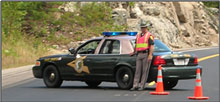 Photo of a New Hampshire State Police cruiser blocking a roadway behind two orange safety cones and a uniformed police officer wearing an orange safety vest.