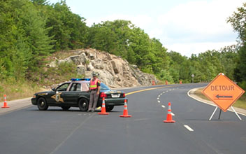 Photo of a New Hampshire State Police cruiser and a uniformed police officer wearing an orange safety vest and blocking a roadway lane behind three orange safety cones and a Detour warning sign that blocks two roadway lanes.